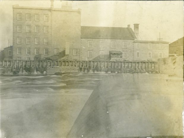 Water flowing over a dam, with a five-storey stone building in the background.