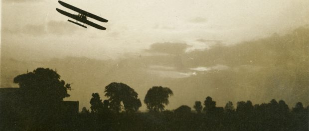 A silhouetted bi-plane flying above trees.