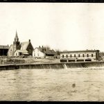 A river with a small dam on it and buildings on the bank behind the dam.