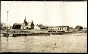 Petit barrage sur une rivière, devant quelques bâtiments. 