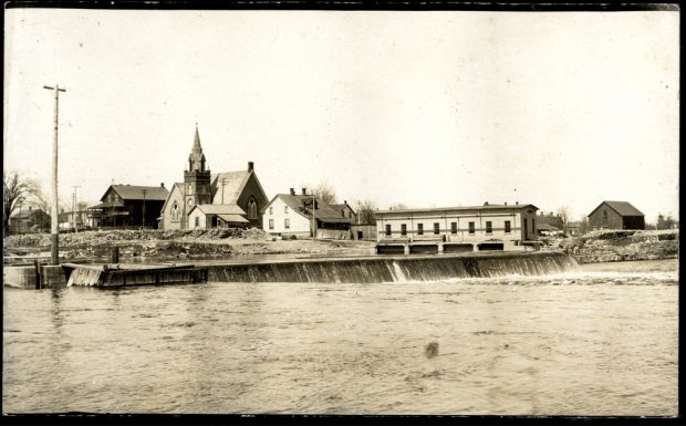 A river with a small dam on it and buildings on the bank behind the dam.