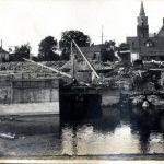 Construction on a river bank and in the river, a church can be seen in the background.