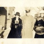 Deux femmes en manteau de fourrure et un homme en uniforme debout dans la neige devant une résidence.