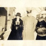 Two women wearing fur coats and a man in military uniform standing in the snow next to a house.