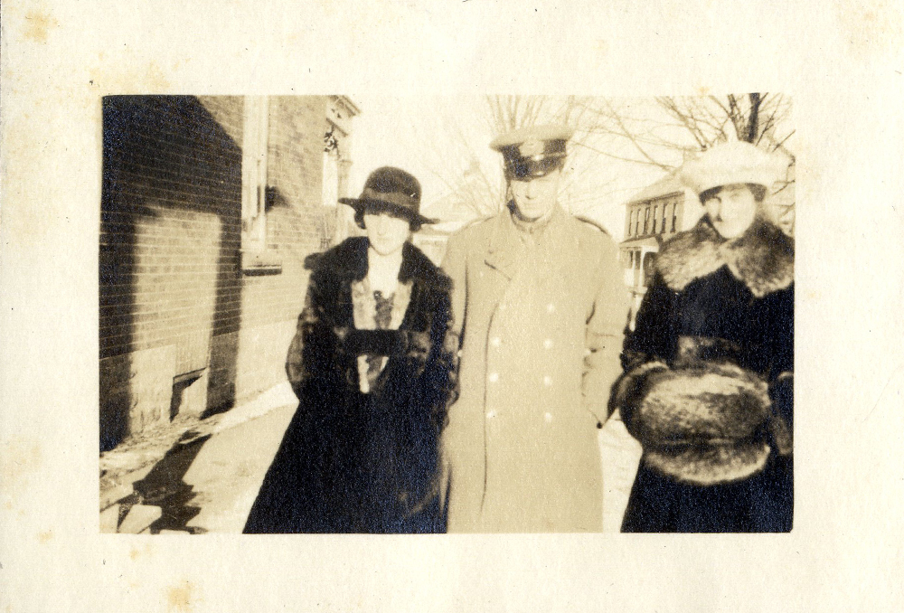 Two women wearing fur coats and a man in military uniform standing in the snow next to a house.