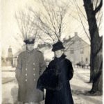 A man in military uniform and woman in a fur coat standing in the snow. Trees and homes in the background.