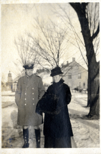 A man in military uniform and woman in a fur coat standing in the snow. Trees and homes in the background.