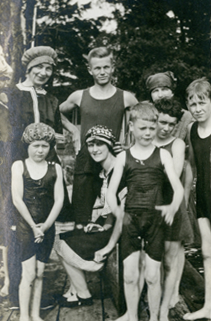 A group of mostly children standing on a dock in bathing suits.