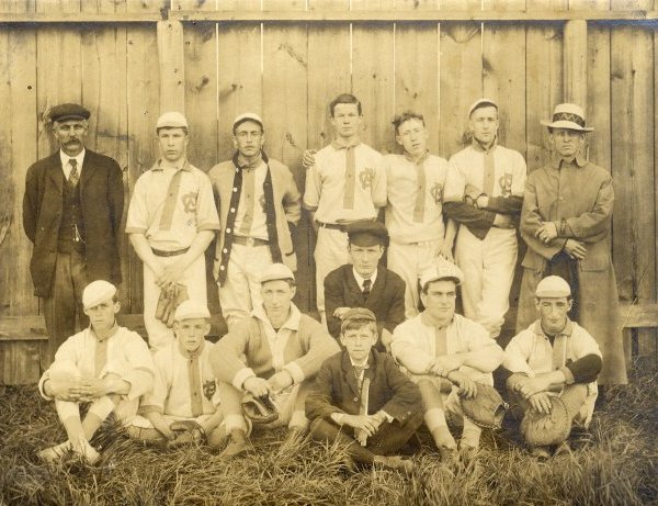 Fourteen men wearing sports uniforms grouped in two rows in front of a wooden fence.