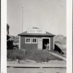 A small single story wood shingled building in the distance. Hand painted General Airways Ltd sign on roof, uncut front lawn, wood board side walk and a man sitting in the doorway, reading.