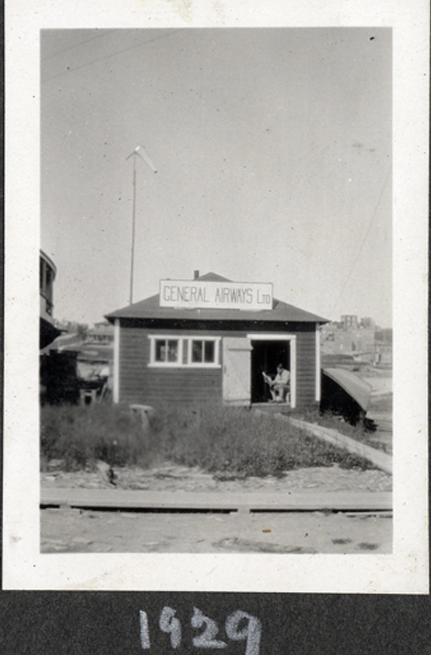 A small single story wood shingled building in the distance. Hand painted General Airways Ltd sign on roof, uncut front lawn, wood board side walk and a man sitting in the doorway, reading.