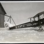 Two pilots dressed in flying gear in front of an open cockpit plane of a bi-plane on skis, in winter, in front of a hangar.