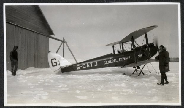 Two pilots dressed in flying gear in front of an open cockpit plane of a bi-plane on skis, in winter, in front of a hangar.