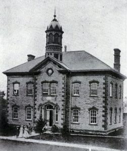 A large, two-storey stone building with a bell tower on the roof. Several women in long dresses are walking out of the front doors.