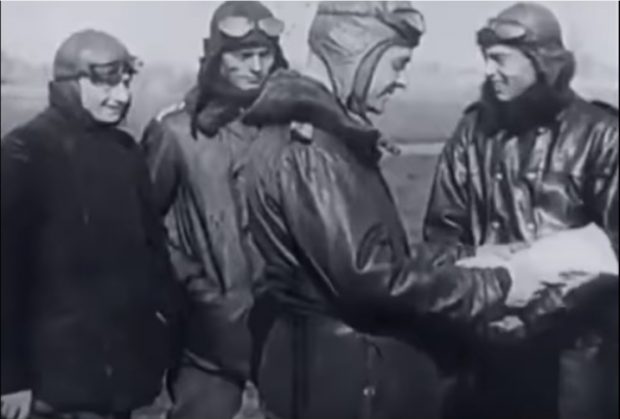 Four men in flying leathers looking at some papers that are being held by one of the men.