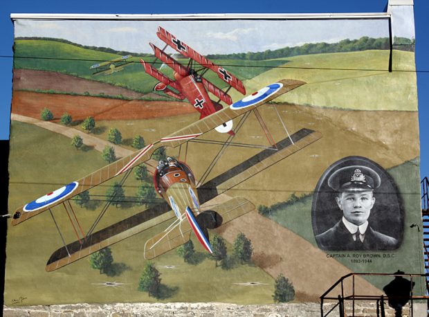 Illustration of two British First World War bi-planes and a German tri-plane in a dog fight over open fields with a man's portrait in the lower right corner.