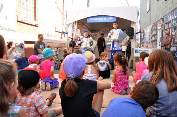 Colour photograph of a group of children watching two young Russian musicians of roughly the same age. The musicians are wearing traditional blue and white Russian clothing.