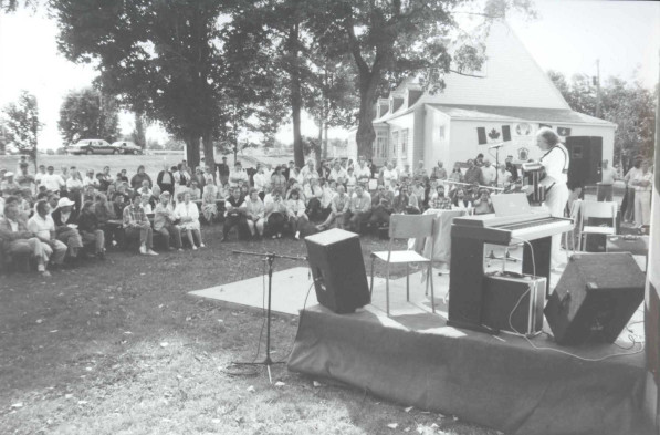 Black and white photograph of a crowd sitting in the shade and listening to Jacques Dumont play the accordion.
