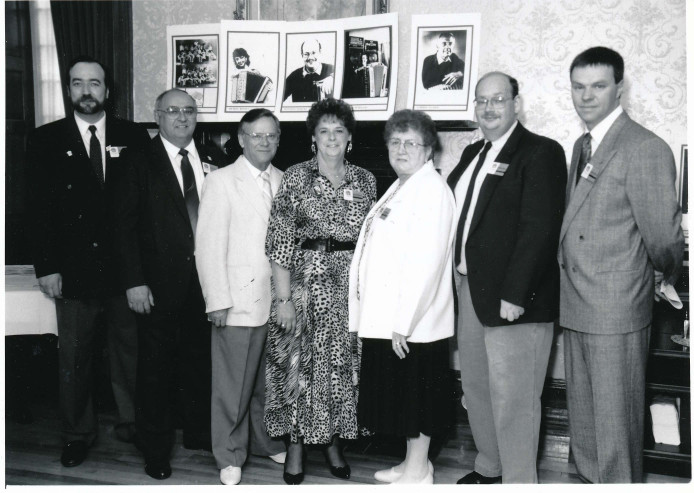 Black and white photograph with several people standing in the foreground. Photographs of artists playing at that year’s Carrefour mondial de l’accordéon appear in the background.