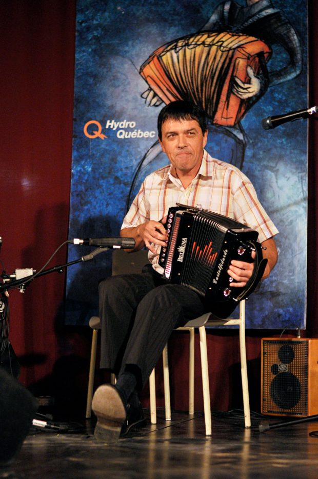 Colour photograph of Denis Pépin sitting on a chair while he plays the diatonic accordion.