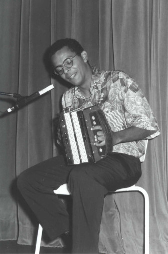 Black and white photograph of Gregory Charles sitting on a chair in front of a stage curtain, holding a diatonic accordion on his knee.