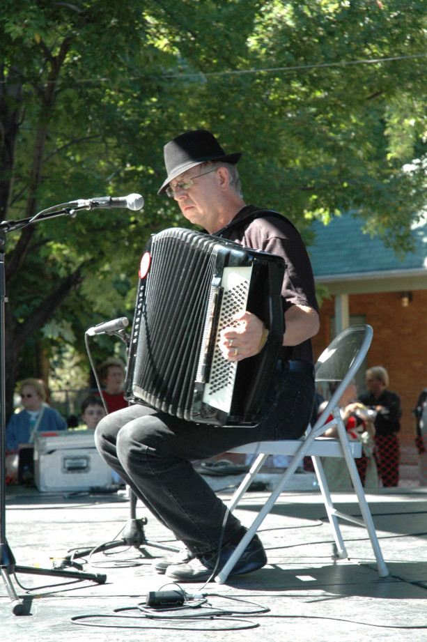 Colour photograph of Len Wallace on an outdoor stage, sitting on a chair while playing his piano accordion.