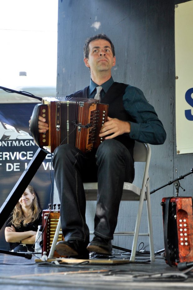 Colour photograph of Sabin Jacques sitting in a chair with his accordion on his knee, tapping his feet to the rhythm of the music as he plays.