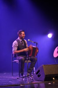 Colour photograph of a smiling Timi Turmel sitting on a chair with a diatonic accordion on his knee.
