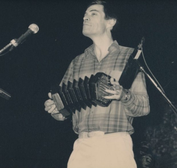 Black and white photography, Alistair Anderson concertina in his hand, plays on an indoor stage.