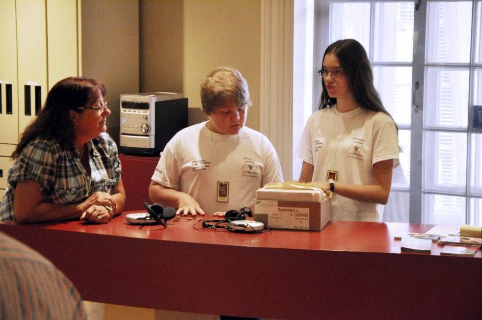 Colour photograph of young volunteers answering a woman’s questions while standing behind a counter at the Musée de l’accordéon.