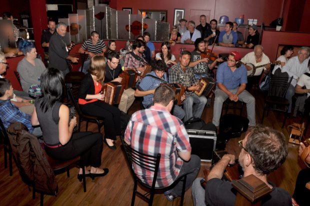 Colour photograph of a large gathering of accordionists and other musicians playing together. They are surrounded by spectators.