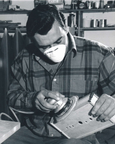 Black and white photograph of Sylvain Vézina sanding an accordion in his workshop. His tools and materials are visible in the background.