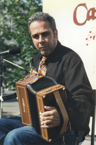 Colour photograph of Alain Pennec sitting on a chair and looking at the camera with an accordion on his knee.