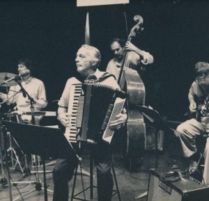 Photographie noir et blanc, Gordon Fleming assis sur un tabouret avec son accordéon-piano, entouré des musiciens qui l’accompagnent.