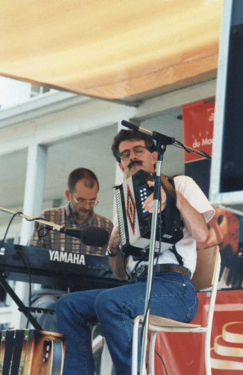 Colour photograph of Gaston Nolet sitting on a chair while holding his diatonic accordion. Luc Lavallée is playing piano in the background.