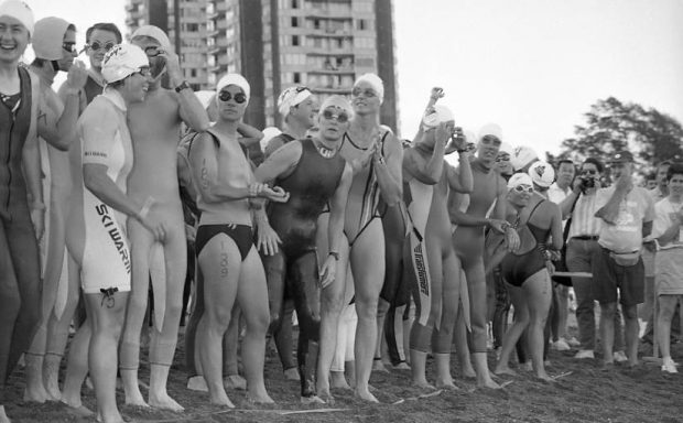Black and white photo of women Triathlon athletes in swim suits, googles, and bathing caps, crowded together at the start line on the beach at English Bay preparing for the 1.5K swim portion of the event.