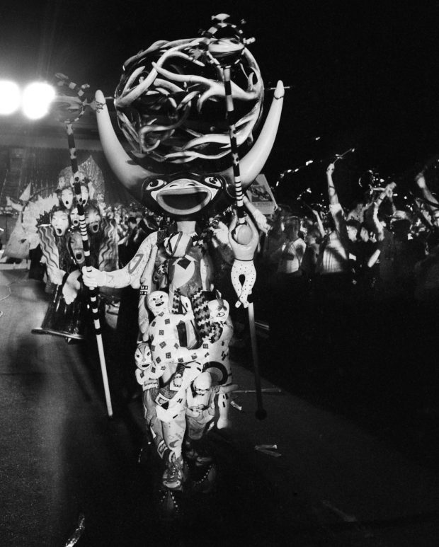 Black and white photo of a character in the Carnaval! fantasy parade. The patterned costume has several three-dimensional baby-sized fabric dolls attached to it. The character has a decorated staff in each hand. One has a baby-sized wood doll clinging to it. The character wears a glossy, painted, wooden mask. It has a wide, open smile and long horns with a large orb-shaped 'crown' comprised of intertwining curved branch-like structures between them.