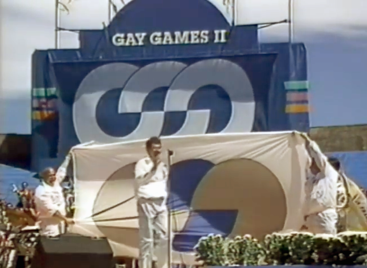 At the Closing Ceremonies of Gay Games II, Richard Dopson stands on the stage at the microphone in front of a large Gay Games flag held out by the Vancouver delegation.