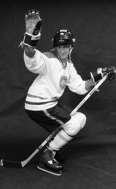 Black and white studio photograph of a male hockey player wearing a Montreal Canadiens white jersey and smiling flirtatiously while posing as if 'riding' his stick.