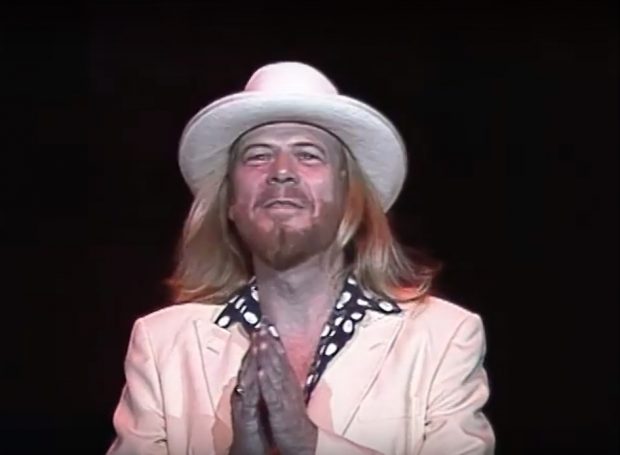 Singer Long John Baldry, wearing a white suit, black shirt with white dots, and white fedora, walks across the stage to the microphone mouthing thank you to the crowd. His band is playing the opening bars of A Thrill's A Thrill.