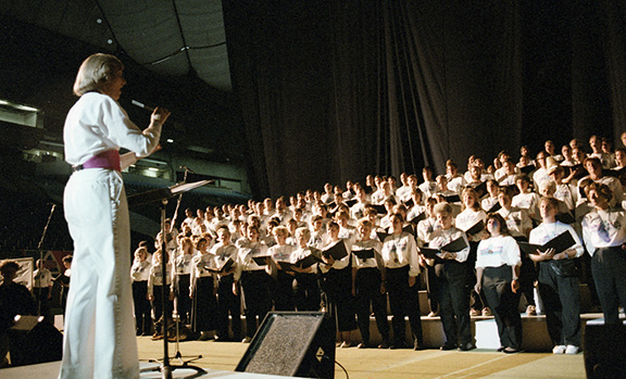 Over 70 members of the Celebration '90 Festival Chorus, in uniform black pants and white shirts, are assembled on a tiered riser on the stage at B.C. Place Stadium. Carole White conducts their vocal performance at the Closing Ceremonies.