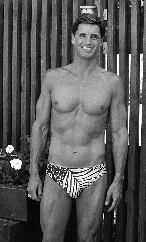 Black and white photo portrait of swimmer Michael Mealiffe. He is wearing swimming trunks and standing on an outdoor patio with a slat fence as background.