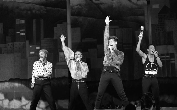 A black and white photo of the four male members of the Canadian a cappella group, The Nylons, striking a pose - wide leg stance, mic in one hand, and the other held straight up towards the roof - as they perform on the B.C. Place Stadium stage for Closing Ceremonies.