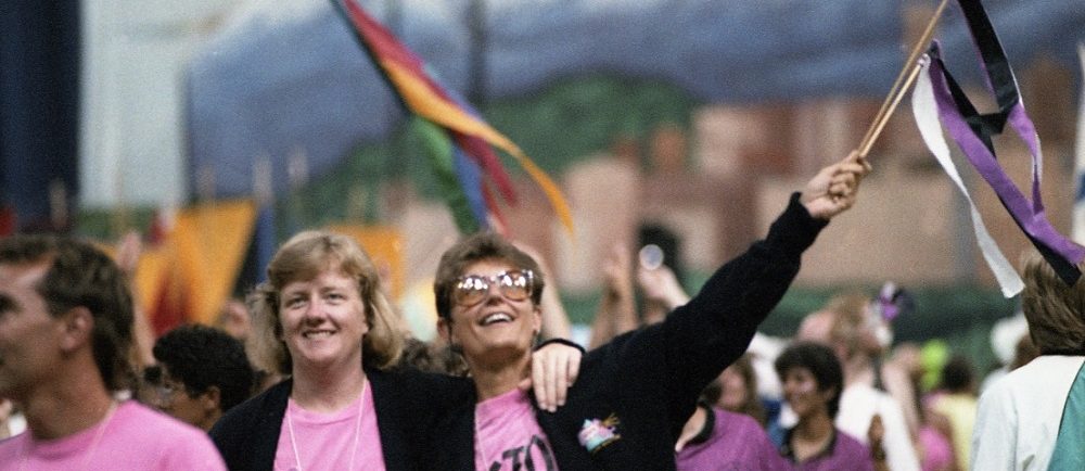 A group of female athletes dressed in pink t-shirts and black jackets smile and wave to the crowd with colourful streamers as they walk in the parade of athletes at Opening Ceremonies. The gigantic mural of Vancouver that backed the stage is visible in the background.