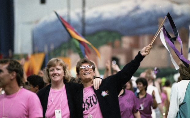 A group of female athletes dressed in pink t-shirts and black jackets smile and wave to the crowd with colourful streamers as they walk in the parade of athletes at Opening Ceremonies. The gigantic mural of Vancouver that backed the stage is visible in the background.