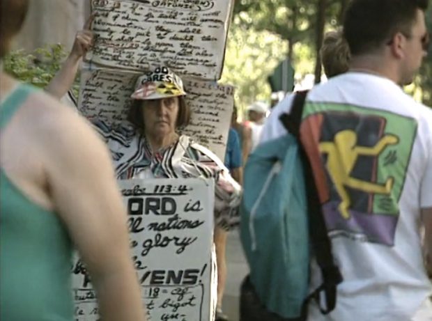 A woman wearing several poster boards with hand-written biblical passages and homophobic rants on them moves through a crowd of Gay Games registrants.