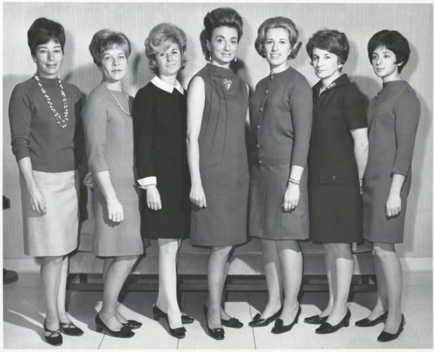A black-and-white formal photograph of a group of women in 1960s-style attire.