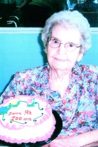 Une femme aux cheveux blancs bouclés portant des lunettes, une chemise fleurie et un collier de perles est assise. Devant elle se trouve un gâteau au glaçage principalement blanc et rose. Sur celui-ci est inscrit : « Bonne fête, 100 ans ».