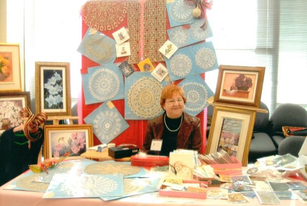 A smiling woman sits behind a table. Behind her, crocheted centrepieces, greeting cards and paintings are displayed. In front of her, crocheted items and cards are arranged on a table.