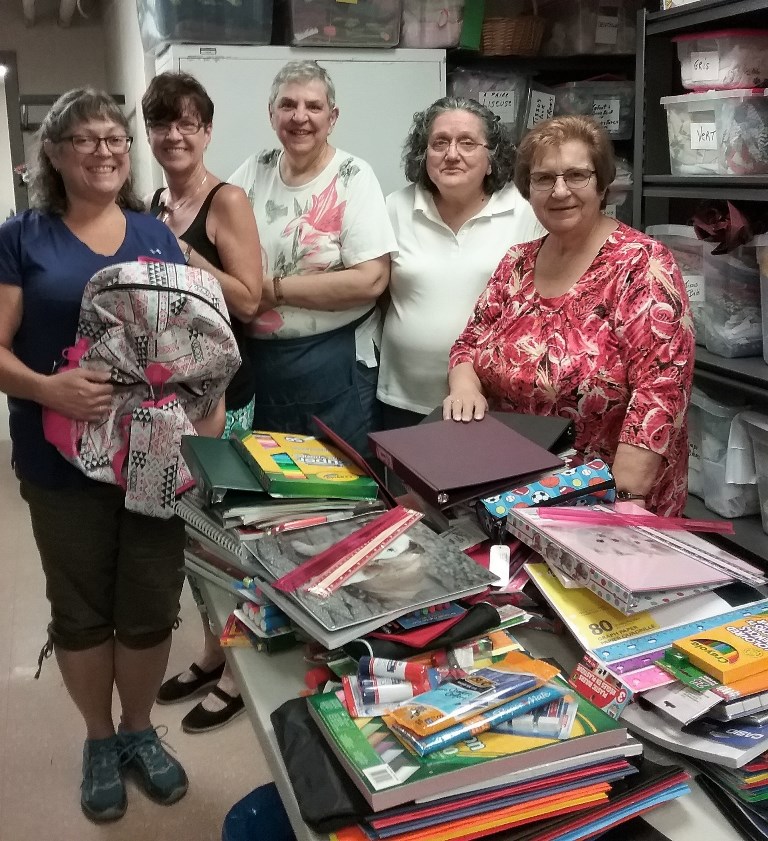 Five smiling women are standing at the end of a rectangular table that is completely covered with various school supplies such as duo-tangs, backpacks and pencils.